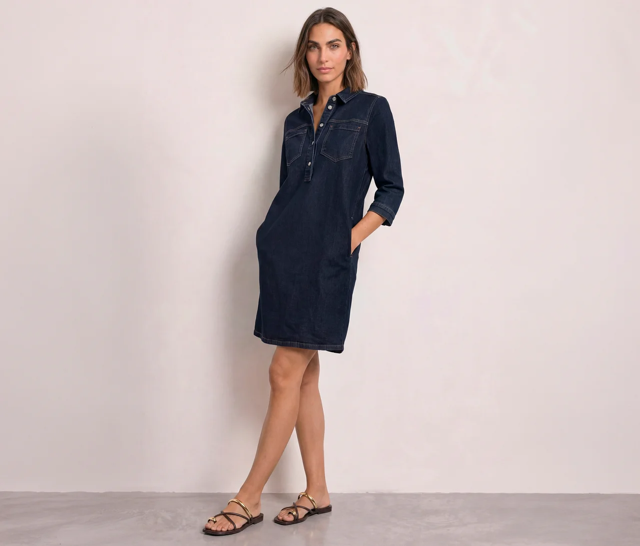 Eine Frau mit kurzen braunen Haaren trägt ein dunkelblaues Street One Kleid aus Denim mit Kragen, Knöpfen, Brusttaschen und Dreiviertelärmeln. Sie steht mit den Händen in den Taschen und trägt dunkle Sandalen mit goldenen Riemen.