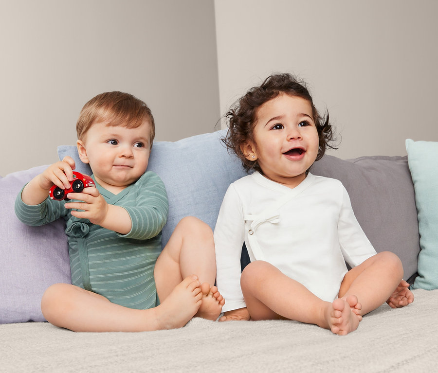 Zwei Babys sitzen auf einem Bett, eines spielt mit einem roten Spielzeugauto, beide tragen 2 Baumwoll-Langarmbodys.