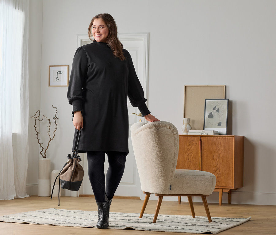 Eine Frau posiert in schwarzen Leder-Stiefeletten, geprägt, einem Sweatkleid und einer Trendstrumpfhose. Sie hat eine Umhängetasche dabei.