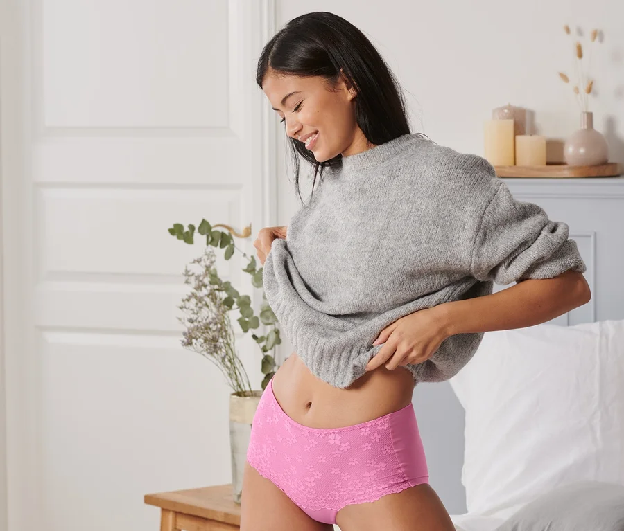 Eine Frau sitzt auf einem Bett und zieht sich einen grauen Pullover über den Kopf. Sie trägt eine pinke Highwaist-Panty.