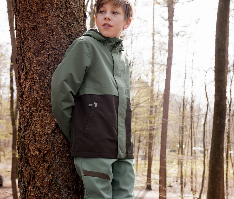 Ein Junge lehnt an einem Baum, er trägt eine Allwetterjacke, eine Softshell-Hose und braune recycelbare Gummistiefel.