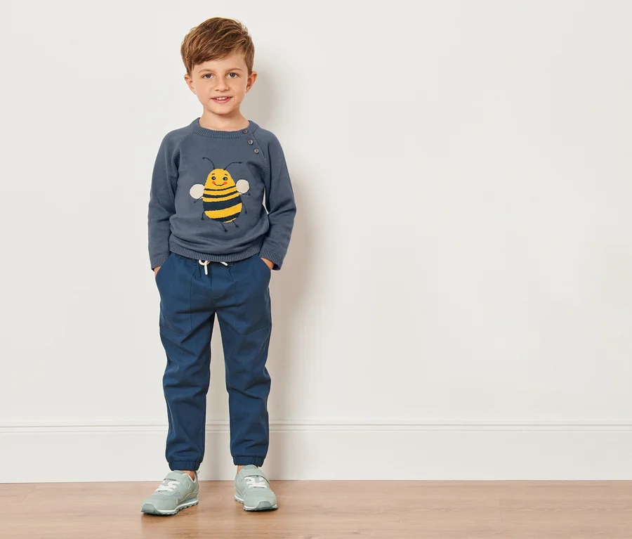 Ein Junge in einem Kleinkinder-Feinstrickpullover mit Bienenmotiv steht vor einer weißen Wand.