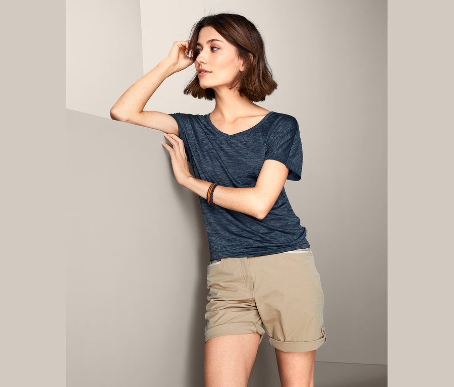 Eine Frau, die sich an eine Wand lehnt, trägt ein dunkelblau meliertes Funktionsshirt und beigefarbene Shorts.