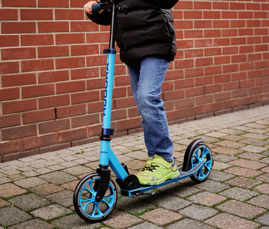 Ein Junge mit Jacke und Helm fährt mit einem blauen HUDORA Up 200 Scooter vor einer Ziegelwand.