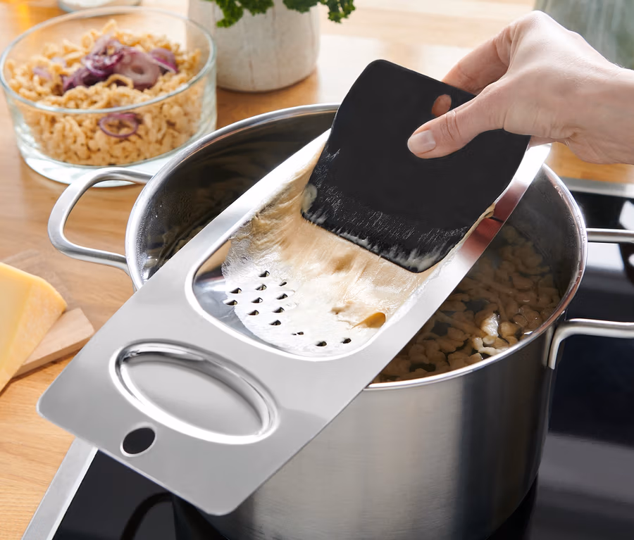 Eine Hand hält eine Spätzlereibe über einen Topf mit kochenden Spätzle.