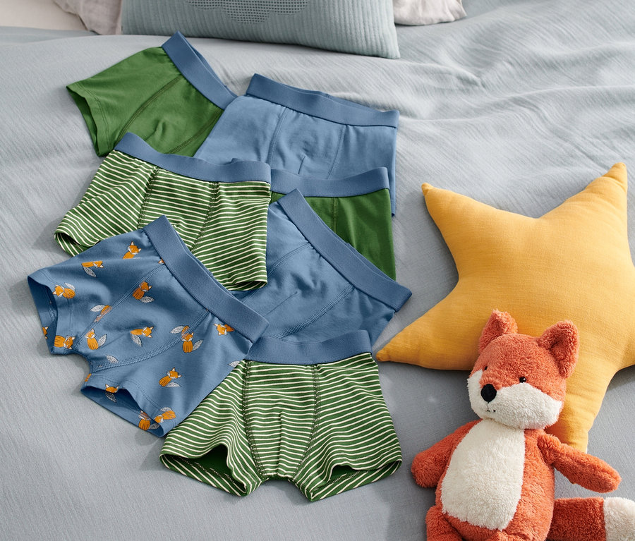 Auf einem Bett liegen sieben Kinder-Slipboxer in verschiedenen Farben und Mustern neben einem Fuchs-Stofftier und einem gelben Stern.