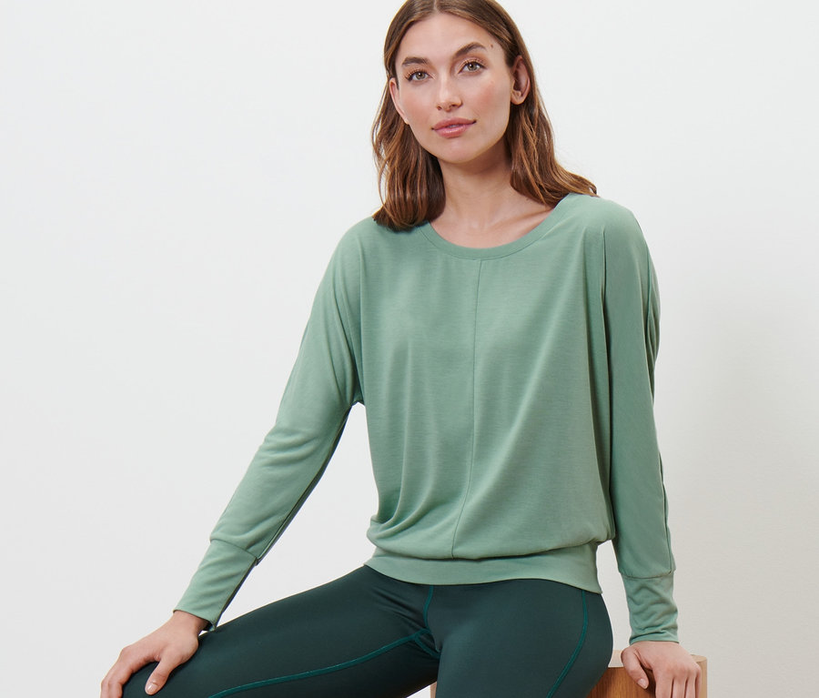 Frau sitzt auf Holzhockern und trägt ein Sport-und-Yogashirt in Salbeigrün.