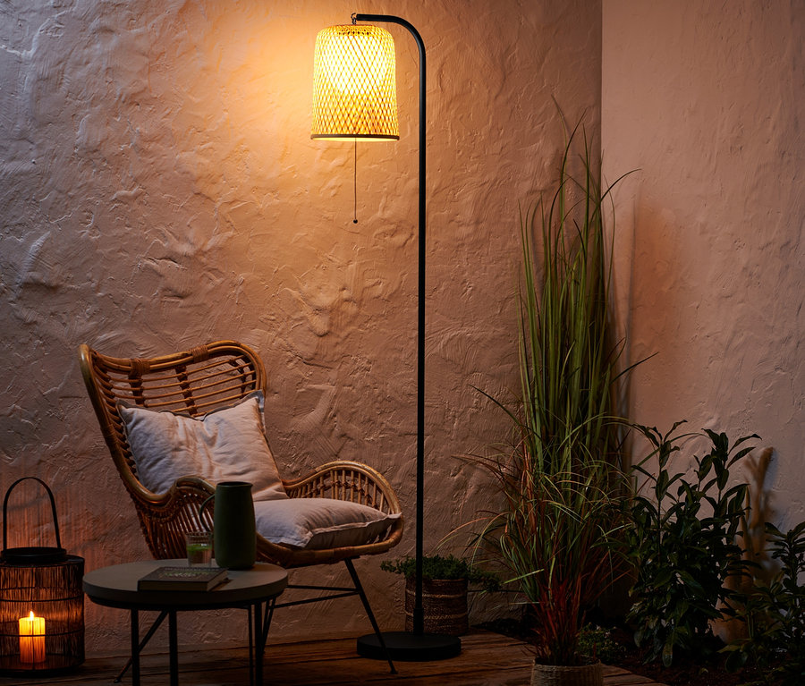 Ein Rattansessel steht neben einem Tisch mit Vase und Buch. Dahinter steht eine Akku-Stehleuchte.