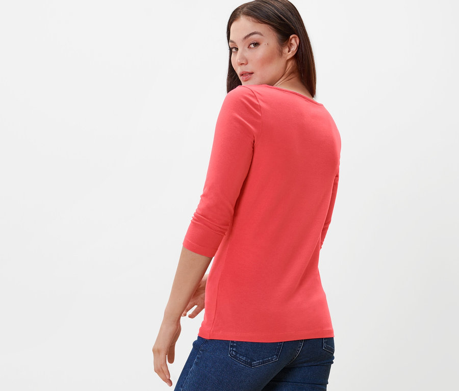 Frau blickt über die Schulter und trägt ein korallefarbenes Shirt mit 3/4-Arm und Jeans.