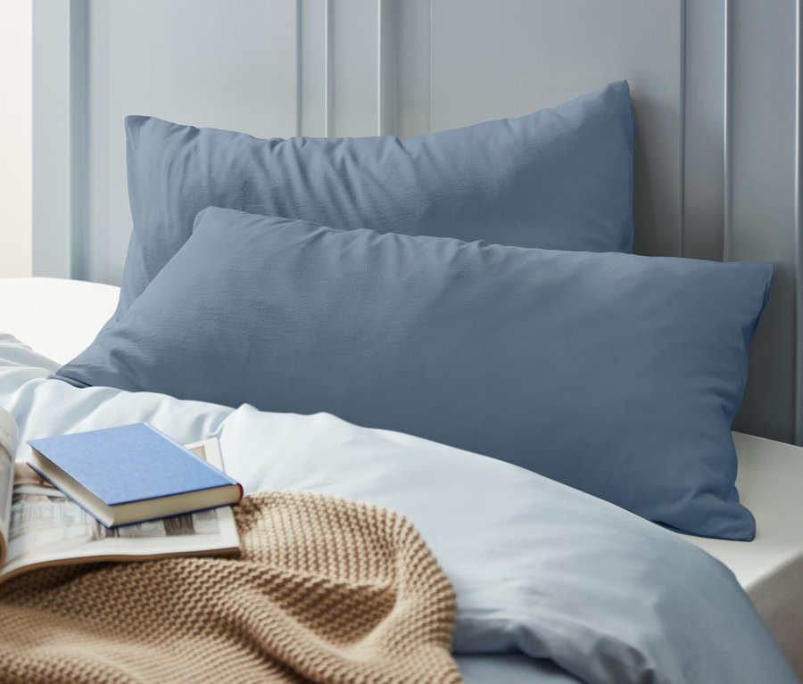 Zwei graublaue Jersey-Kopfkissenbezüge liegen hintereinander auf einem Bett. Auf der Decke liegen ein aufgeschlagenes Buch und ein blaues Buch.
