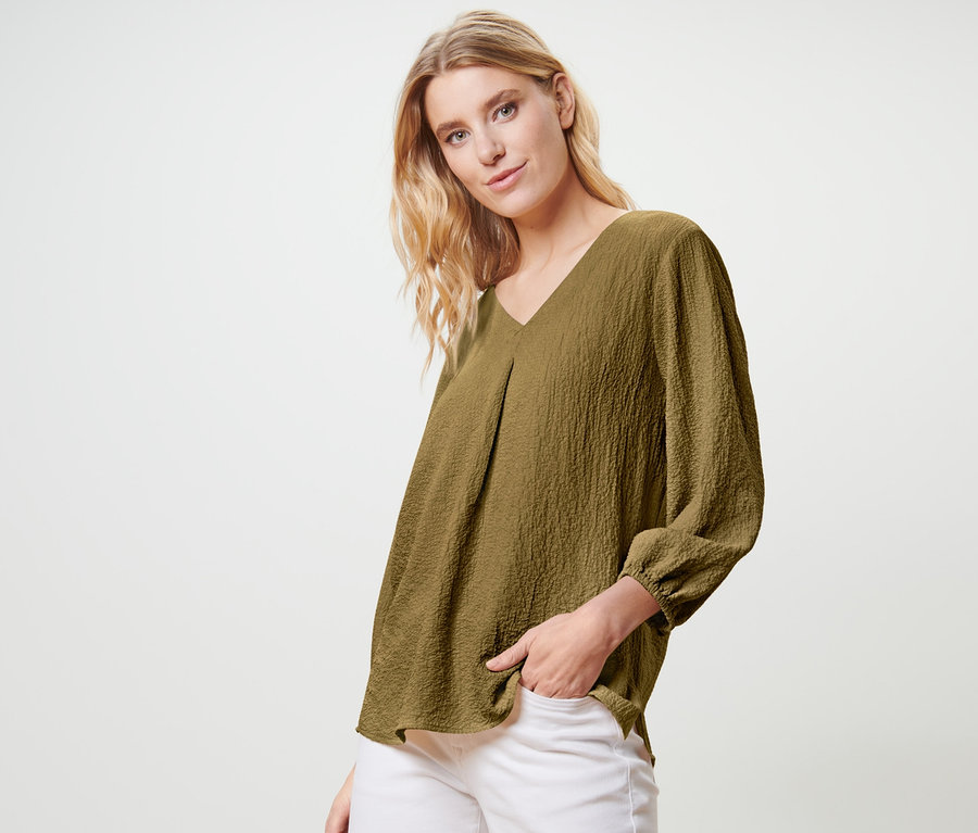 Blonde Frau posiert in olivgrüner Tunika-Bluse und weißen Hosen.