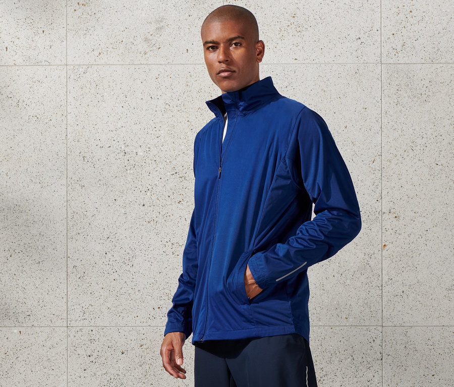 Mann trägt eine blaue Windprotection-Funktionsjacke, Shorts und weiße Turnschuhe.