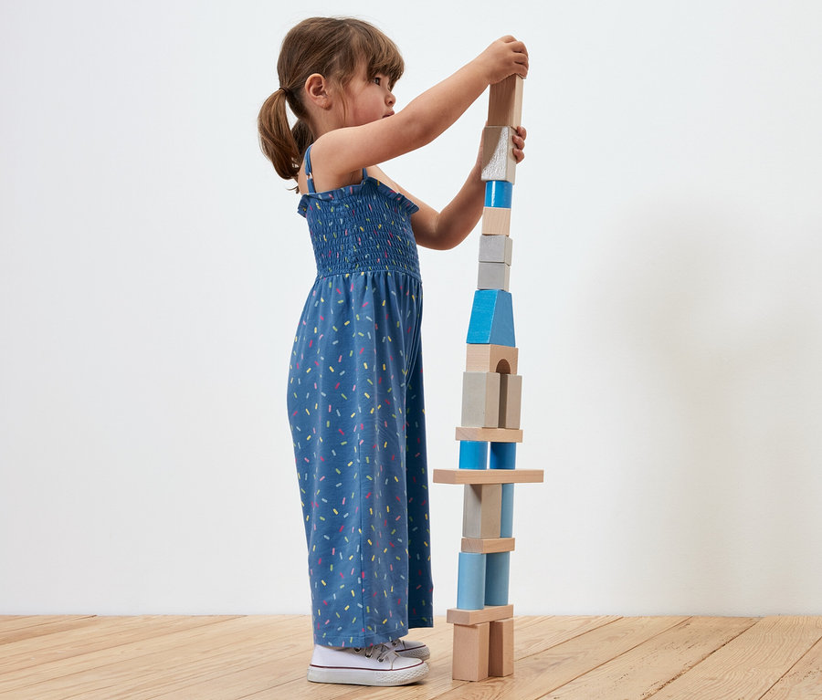 Ein Mädchen baut einen Turm aus Holzklötzen. Sie trägt einen Jumpsuit in 7/8-Länge.