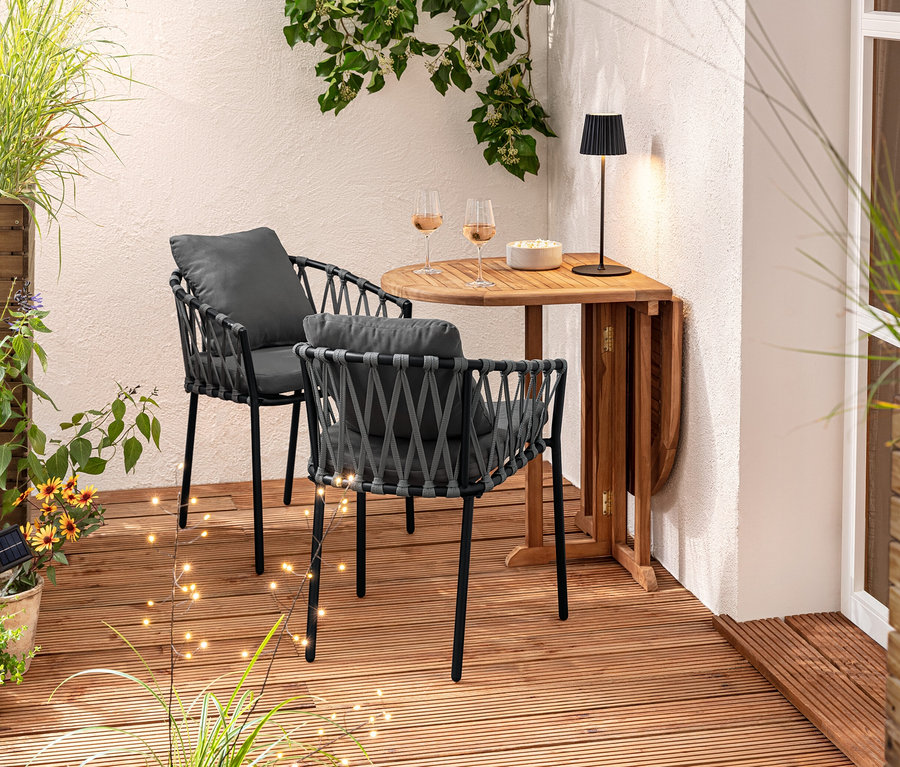 Zwei Dining-Sessel mit Textilgeflecht stehen an einem Holztisch. Auf dem Tisch stehen Gläser und eine Schüssel. Im Hintergrund befindet sich ein Pflanzkasten-Raumteiler und 3 Solar-Spieße.