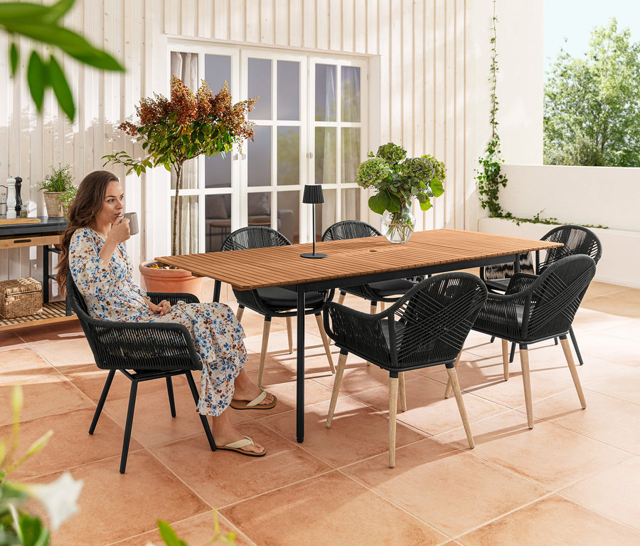 Eine Frau sitzt auf der Terrasse in einem drehbaren Esssessel mit Textilgeflecht und trinkt Kaffee. Im Hintergrund sind ein Ausziehtisch Elin, ein Grillwagen Elin und eine LED-Tischleuchte zu sehen.