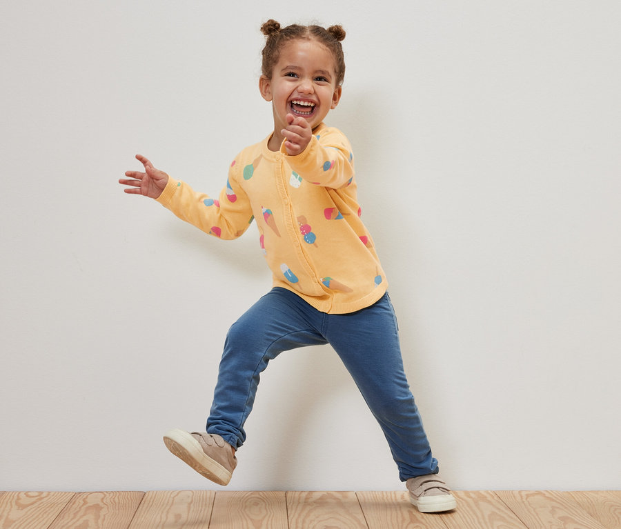 Lächelndes Mädchen springt und trägt eine Kinder-Strickjacke mit Eis-Motiv.