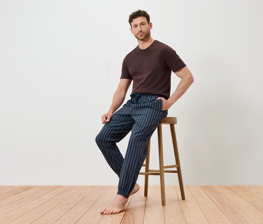 Mann sitzt auf einem Holzhocker, trägt ein braunes T-Shirt und eine blau gestreifte Hose.