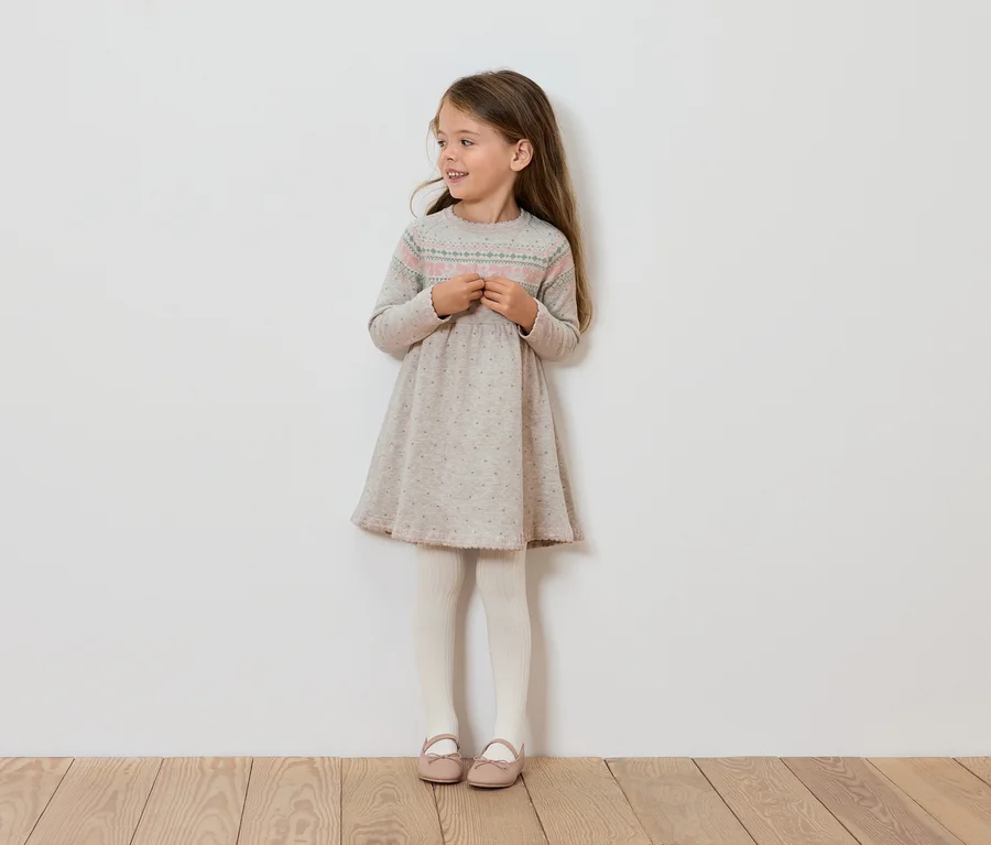 Lächelndes Mädchen in einem langärmeligen Kleid, weißen Leggings und rosa Schuhen steht an einer Wand.