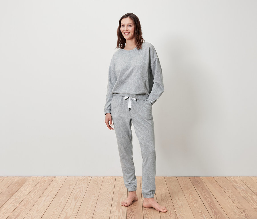 Frau steht und lächelt, sie trägt einen grauen Pullover und eine Jogginghose.