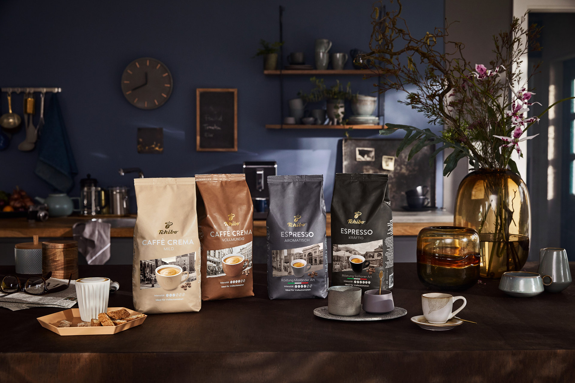 Vier Packungen Tchibo Kaffee stehen auf einem Tisch neben Tassen und Dekorationen.