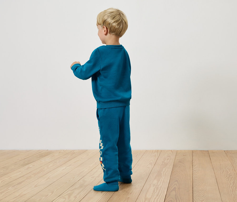 Junge in türkisfarbenem Trainingsanzug steht seitlich auf Holzboden.