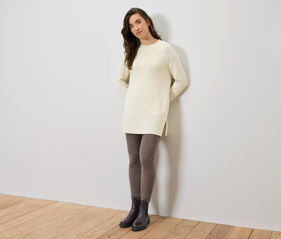 Frau steht und trägt einen langen Strickpullover in Creme, graue Leggings und schwarze Stiefel.