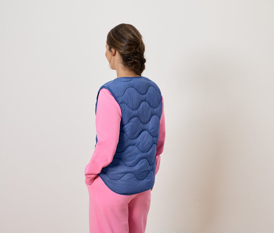 Frau mit dem Rücken zur Kamera trägt eine blaue Steppweste über einem rosa Pyjama.