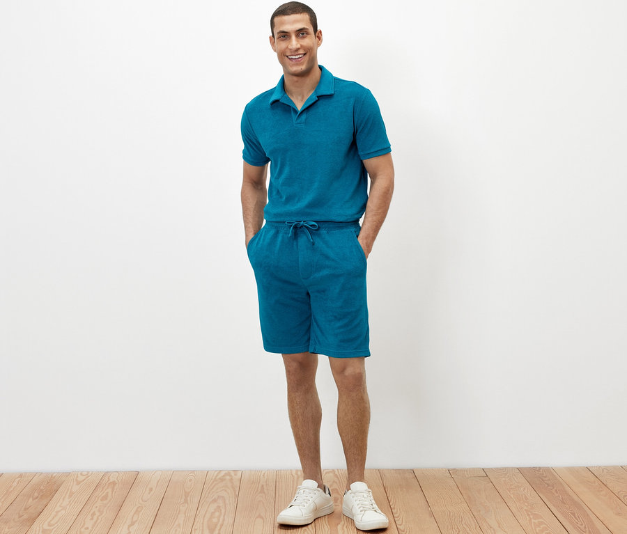 Mann steht mit den Händen in den Taschen seiner petrolfarbenen Frottee-Shorts.