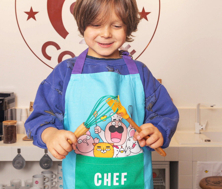 Ein Junge hält einen Schneebesen und einen Spatel aus dem Chefclub Koch-Set und trägt eine Schürze.