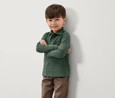 Ein Junge steht mit verschränkten Armen an einer Wand. Er trägt ein grünes Langarm-Poloshirt und braune Hosen.
