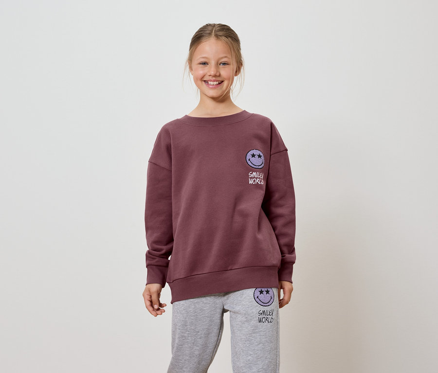 Lächelndes Mädchen trägt ein burgunderfarbenes Sweatshirt und graue Jogginghose mit Smiley World Logo.
