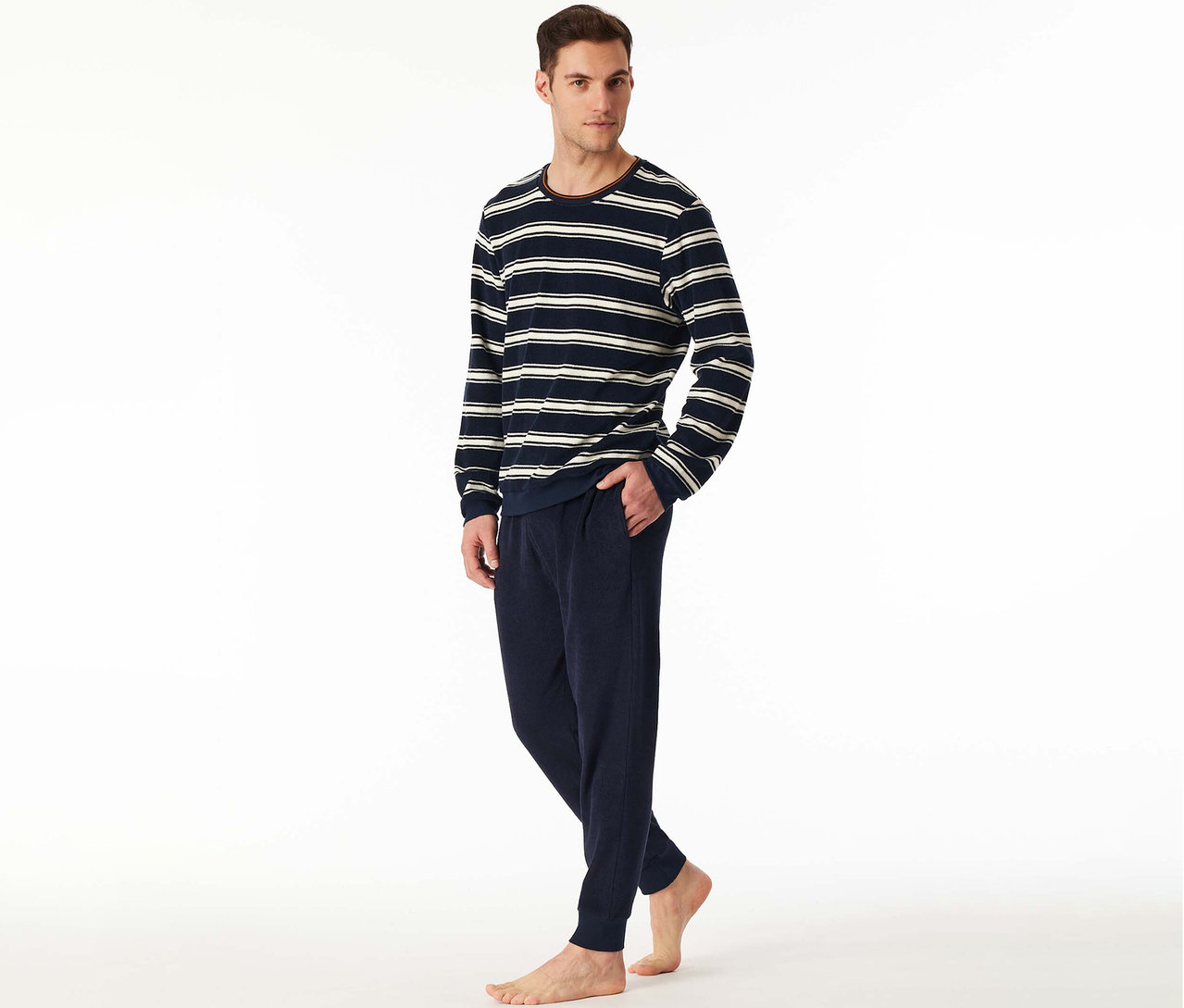 Mann trägt ein SCHIESSER Frottee-Pyjama-Set mit gestreiftem Langarmshirt in Marineblau-Weiß und marineblauer Hose, barfuß stehend.