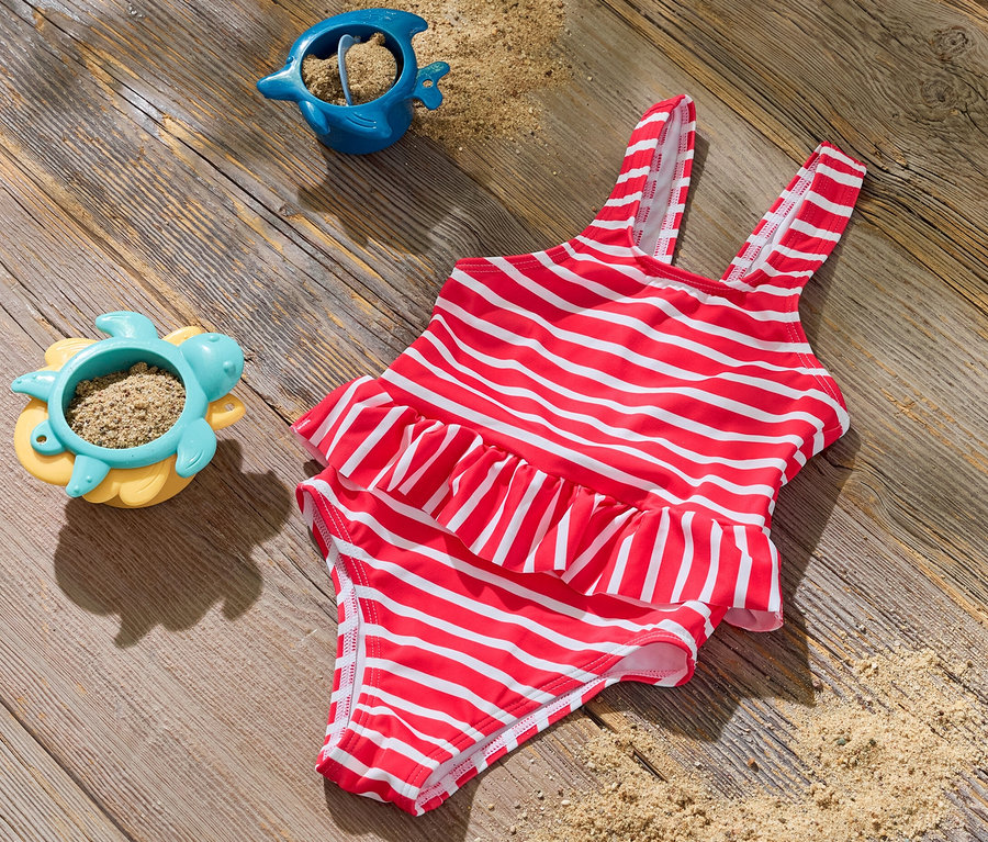 Ein Kinder-Badeanzug mit roten und weißen Streifen liegt auf einer Holzoberfläche neben zwei Sandspielzeugen.