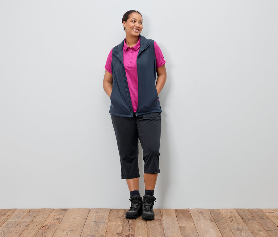 Frau steht mit den Händen in den Taschen vor einer Wand. Sie trägt eine marineblaue Weste über einem pinkfarbenen Poloshirt, dazu eine schwarze Hose und Stiefel.