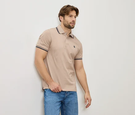 Ein Mann in einem beigen Poloshirt und Jeans steht an einer Wand.