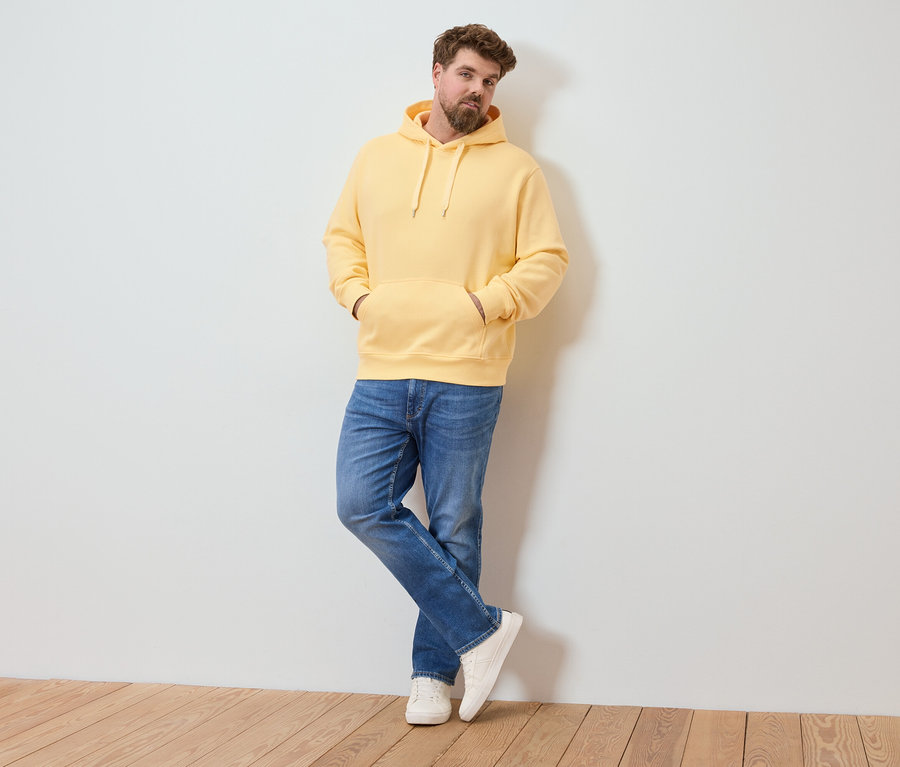 Mann mit Bart lehnt an einer Wand, trägt einen gelben Hoodie, Jeans und weiße Turnschuhe.