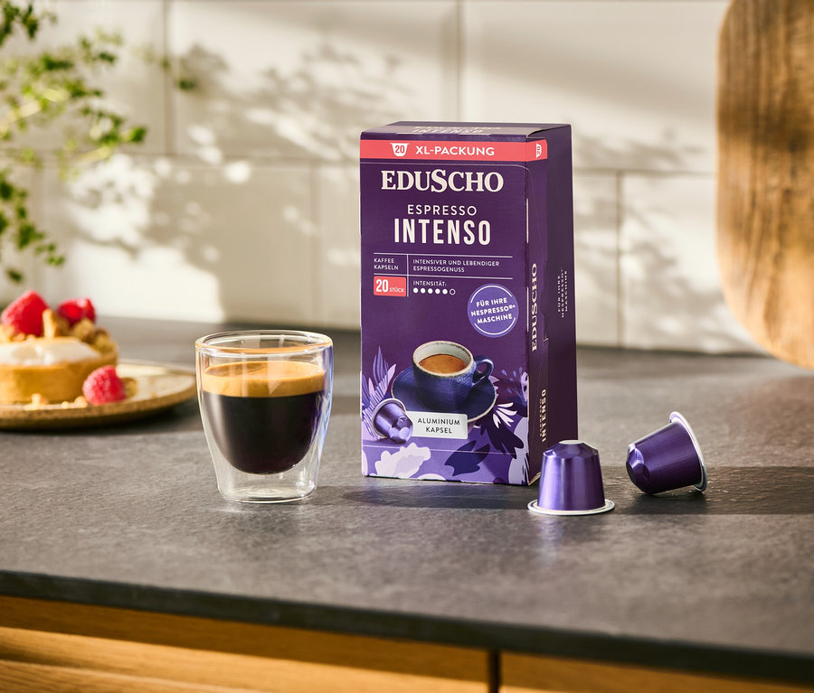 Ein Glas Espresso steht neben einer Packung EDUSCHO Espresso Intenso - 20 Kapseln und zwei Kaffeekapseln auf einer Küchentheke.