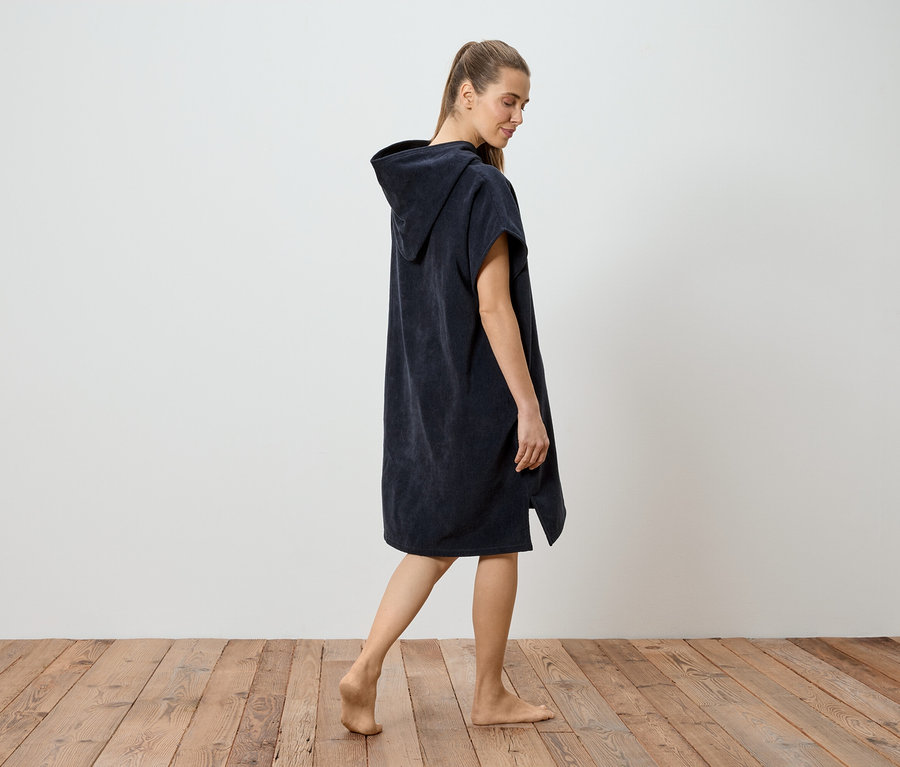 Barfuß geht eine Frau in einem marineblauen Kapuzenponcho über einen Holzboden vor einer weißen Wand.