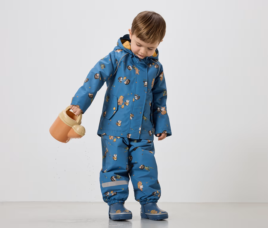 Ein Kind gießt mit einer orangefarbenen Gießkanne Wasser. Es trägt einen blauen Regenanzug mit Tiermotiven und passende Gummistiefel.
