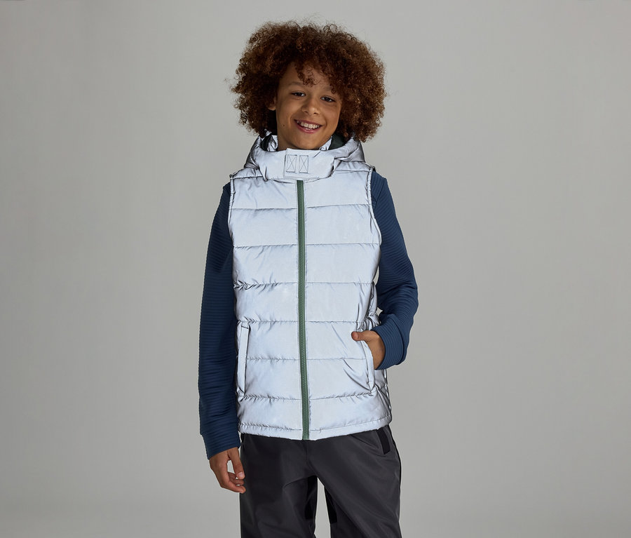Ein Junge mit Afro steht in einer reflektierenden Weste mit Kapuze über einem blauen Pullover und grauen Hosen vor einem grauen Hintergrund.