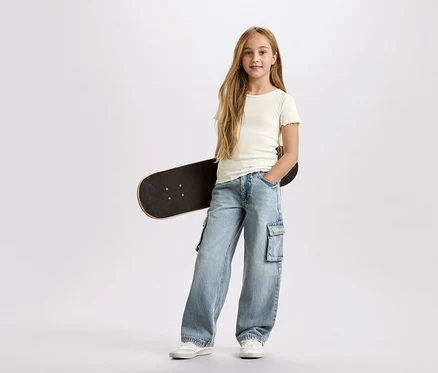 Mädchen mit langen blonden Haaren, mit einem schwarzen Skateboard unter dem Arm, trägt eine hellblaue Cargohose und ein weißes T-Shirt vor einem weißen Hintergrund.