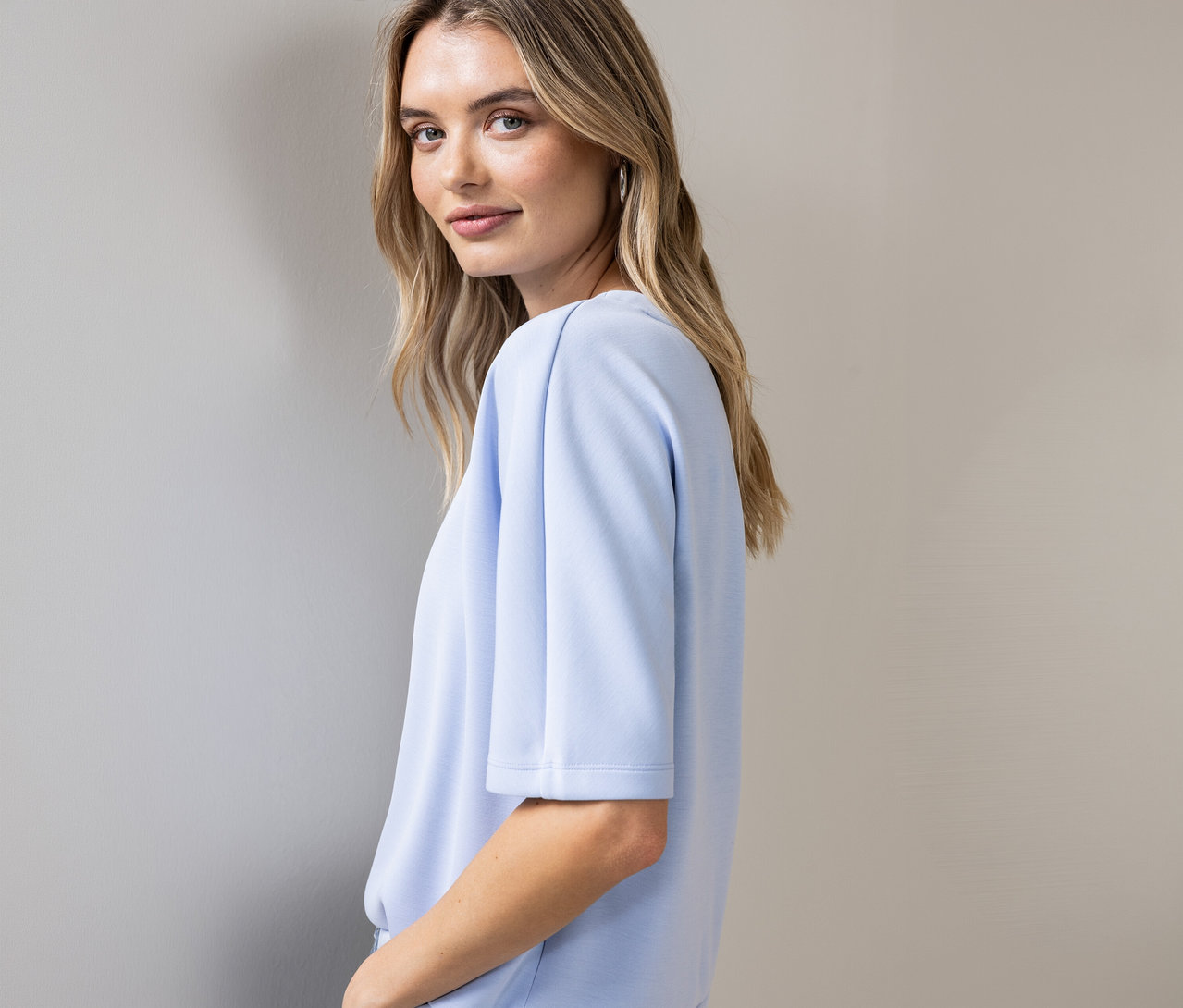 Frau blickt über ihre Schulter und trägt ein hellblaues Street One Shirt mit kurzen Ärmeln und silberne Creolen.