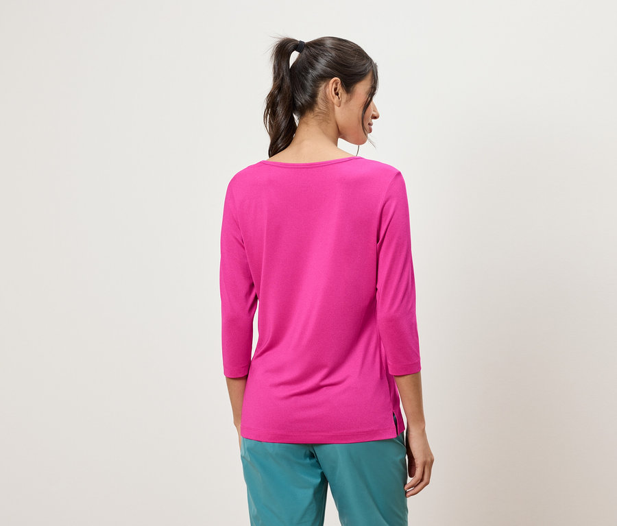 Rückansicht einer Frau mit Pferdeschwanz, die ein pinkfarbenes Dreiviertelarm-Shirt und türkisfarbene Hosen vor einem cremefarbenen Hintergrund trägt.