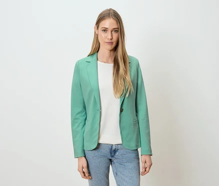Frau trägt grünen Blazer, weißes Shirt und blaue Jeans vor einem weißen Hintergrund.