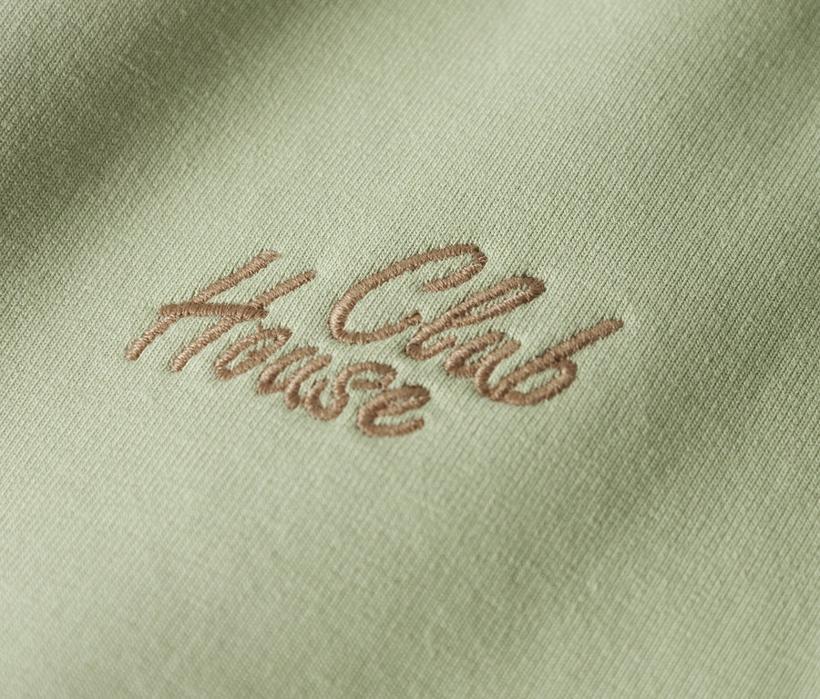 Nahaufnahme eines hellgrünen Stoffes mit der braunen Stickerei "House Club".