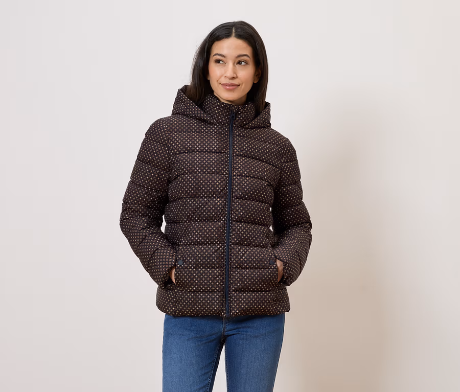 Frau mit brauner, gemusterter Daunenjacke mit Kapuze und blauen Jeans, die mit den Händen in den Taschen vor einer weißen Wand posiert.