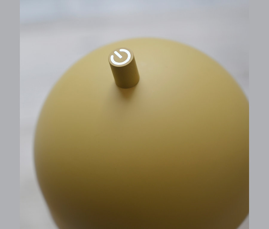 Nahaufnahme eines runden, matten gelben Geräts mit einem kleinen zylindrischen Knopf oben, der ein weißes Einschaltsymbol zeigt.