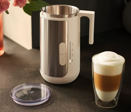 Ein Milchaufschäumer aus Edelstahl steht neben einem Glas Latte Macchiato und einem Deckel auf einer dunklen Arbeitsfläche.
