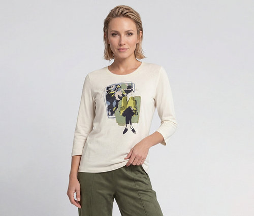 Blonde Frau trägt cremefarbenes Langarmshirt mit grafischem Druck einer Frau in gelber Jacke und olivgrüne Hose.