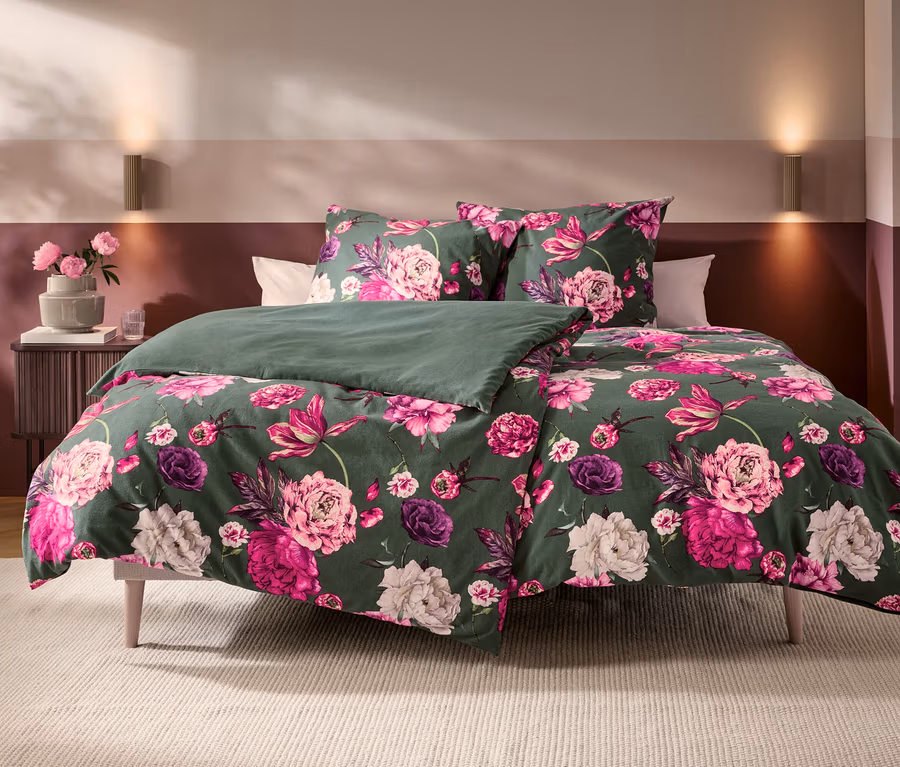 Bett mit dunkelgrüner Bettwäsche, die mit großen rosa und violetten Blumenmustern verziert ist. Eine Seite der Decke ist schlicht dunkelgrün. Daneben steht ein Nachttisch mit rosa Blumen in einer Vase.